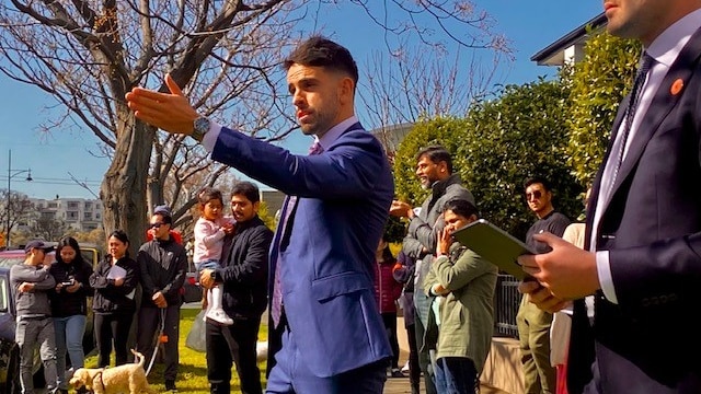 Auctioneer gestures to the crowd in front of a house he is selling. The sky is blue and a large crowd gather to watch