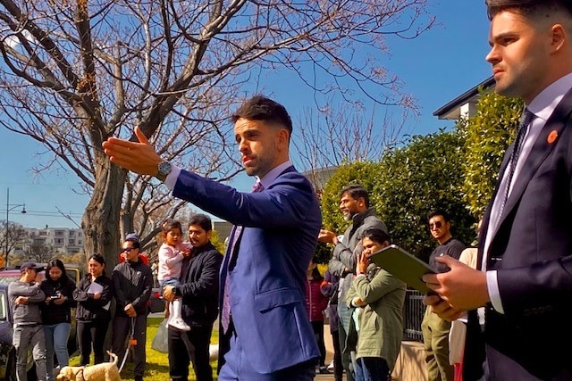 The auctioneer gestures to the crowd outside a house he is selling.  The sky is blue and a large crowd gathers to watch. 