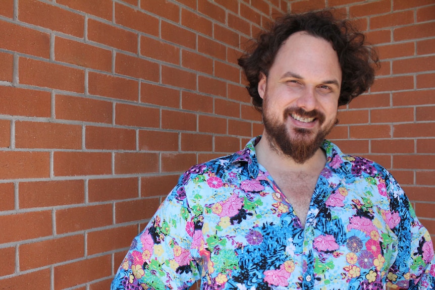 Man wearing a colourful floral shirt smiling in front of a brick wall.