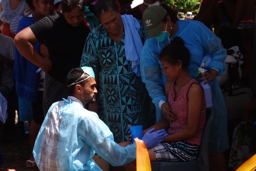 A woman sits while a man in medical scrubs sits in front of her and others line up around them.