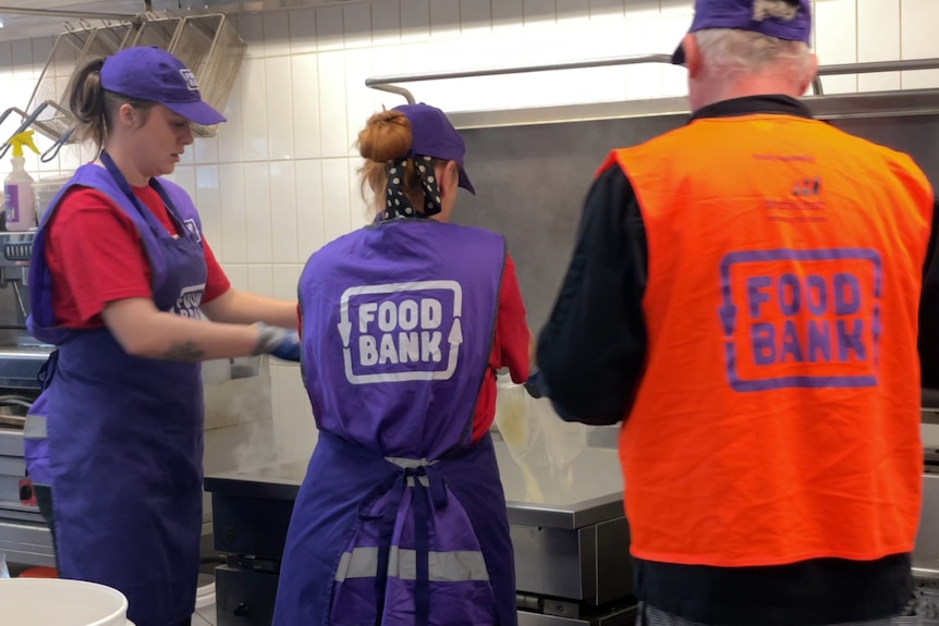 three people in shirts with FOODBANK written on the back stand facing a commercial kitchen 