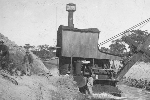 Two men stand on the banks of a creek, while two men walk in the creek next to a large steam-powered shoveling machine.