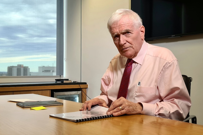 A man in a pink shirt with a red tie sits at a desk looking at papers and into the camera