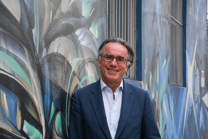 A man with greying hair, wearing a suit jacket and white shirt, stands in front of a colourful wall. 