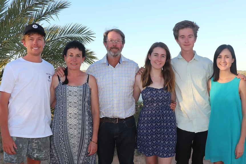 A family smiles at the camera, their arms are around each other and they are standing in front of date palms on a sunny day.