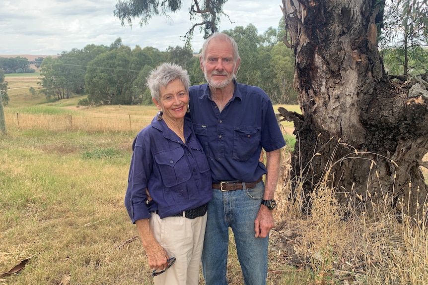 A woman and man stand together with trees and grassland in the background.