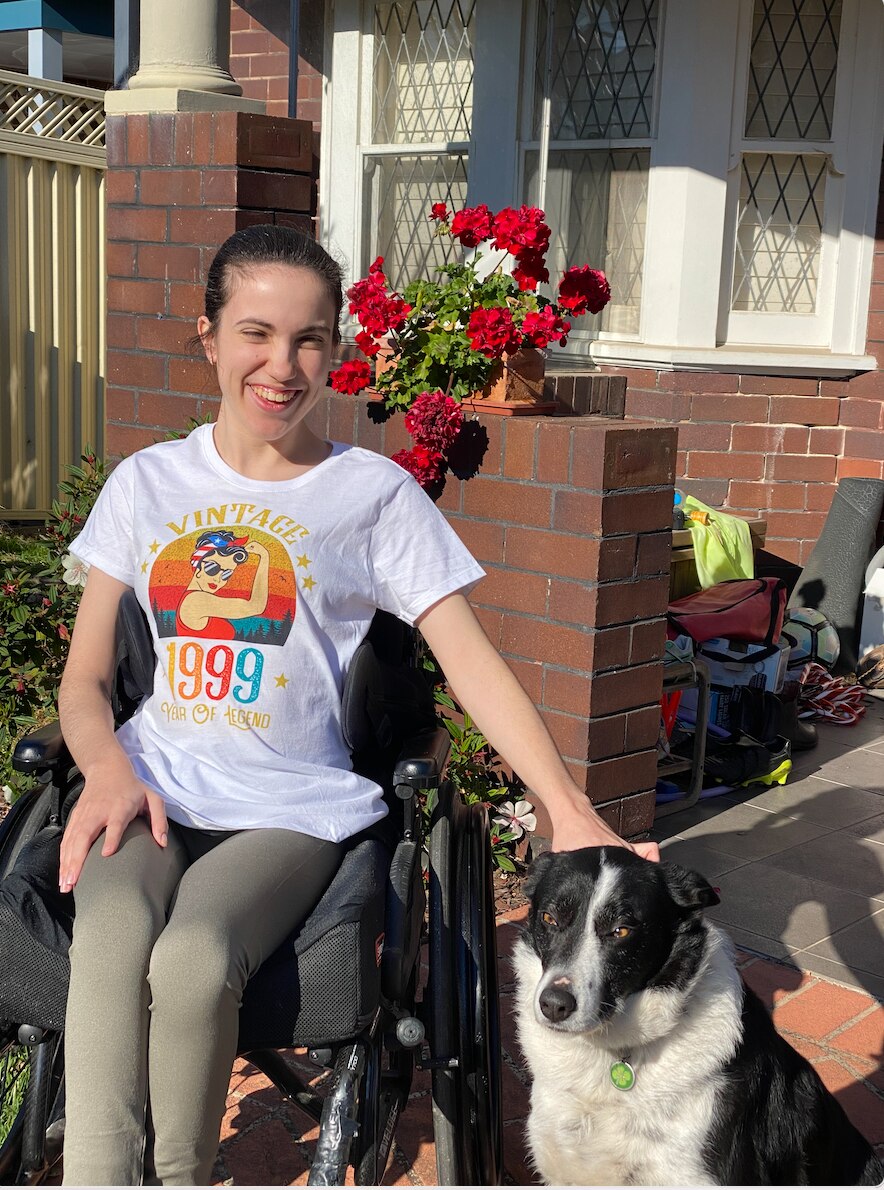Hannah Diviney smiling while sitting outside a house in her wheelchair, patting a dog. Wearing a graphic t shirt and leggings.