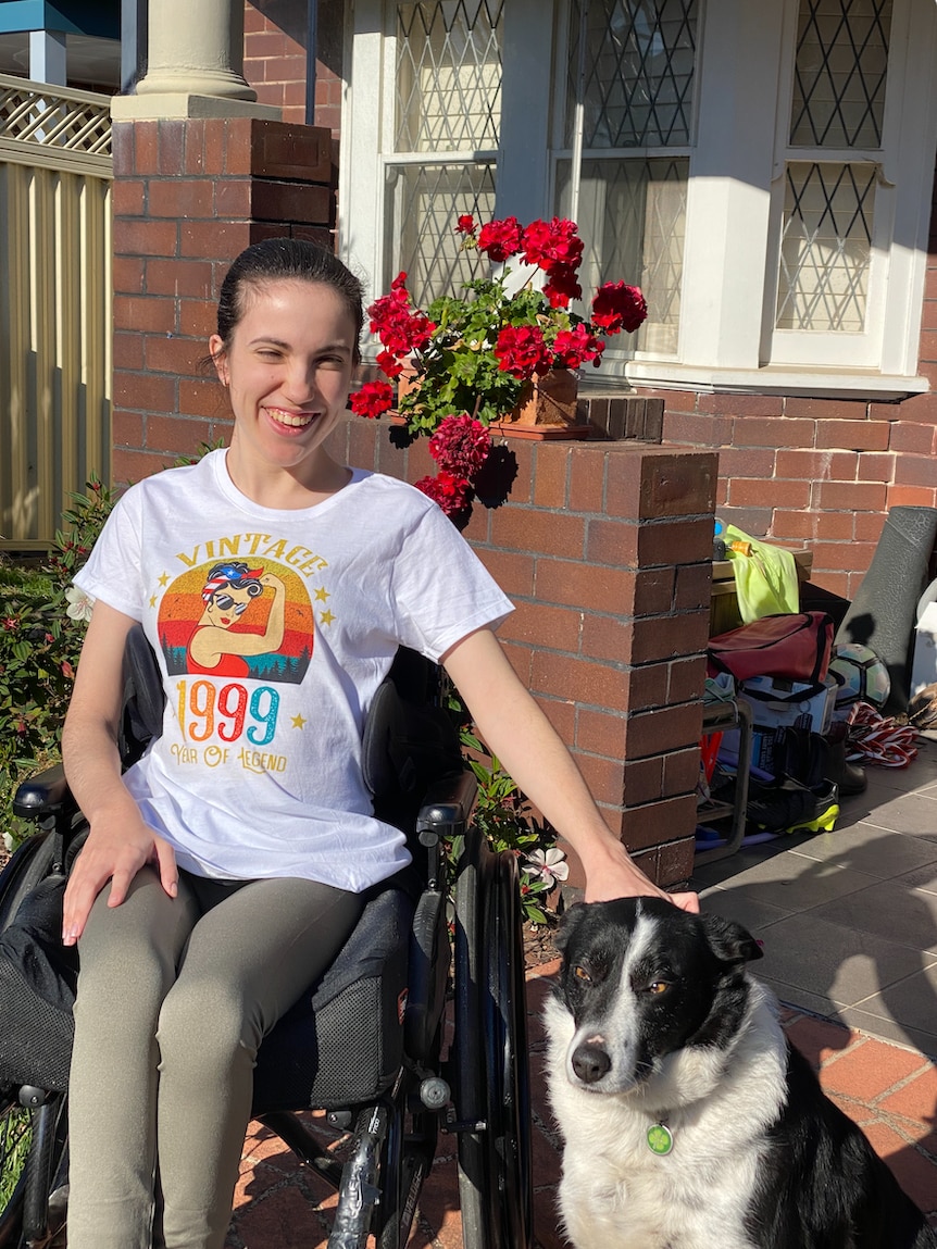 Hannah Diviney smiling while sitting outside a house in her wheelchair, patting a dog. Wearing a graphic t shirt and leggings.