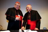 Two older white men wearing black cardinal robes, crosses and red caps.