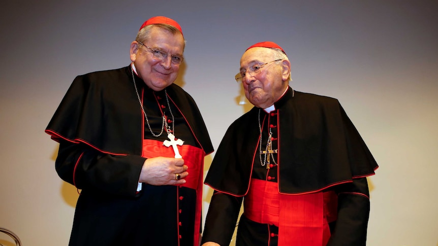 Two older white men wearing black cardinal robes, crosses and red caps.