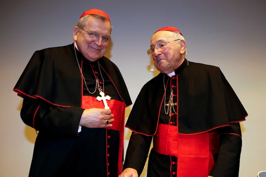 Two older white men wearing black cardinal robes, crosses and red caps.