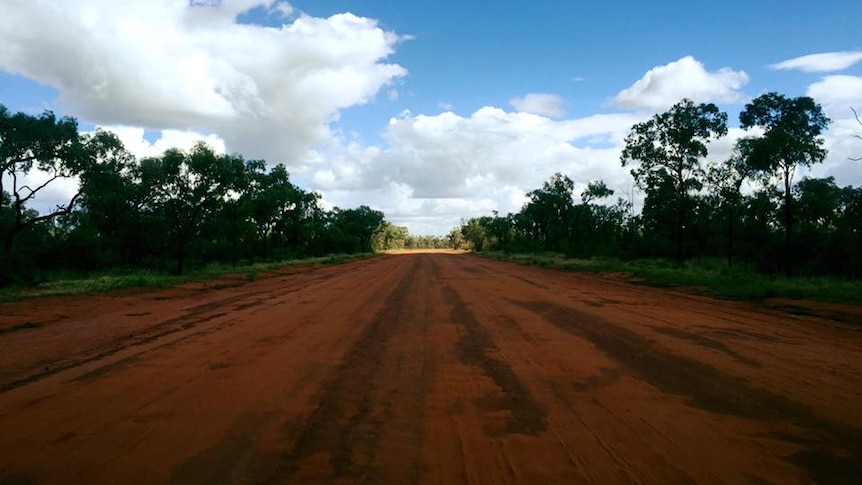 A red dirt road stretches into the distance with blue skies above it