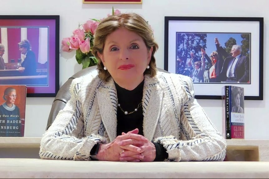 A woman in a white jacket sits at a desk with her hands clasped in front of her.