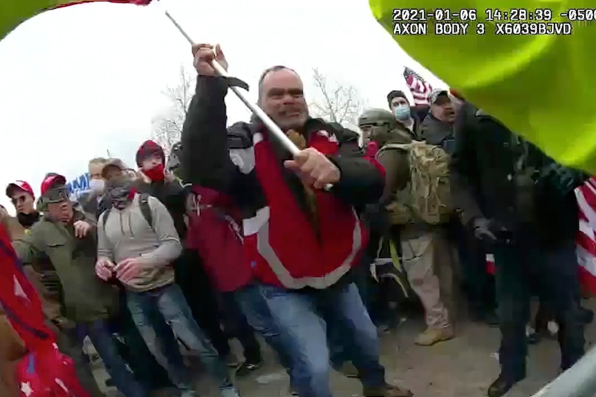 A man in a red jacked holds up a metal pole and is directing it towards the camera. 