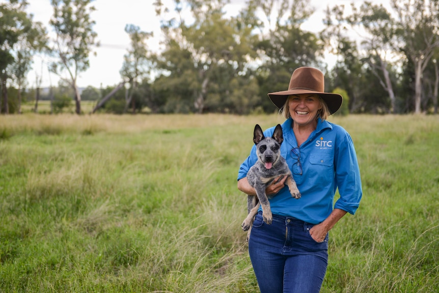 A woman in a blue button-up shirt and wide brim hat stands in a grass field smiling while holding a blue heeler puppy