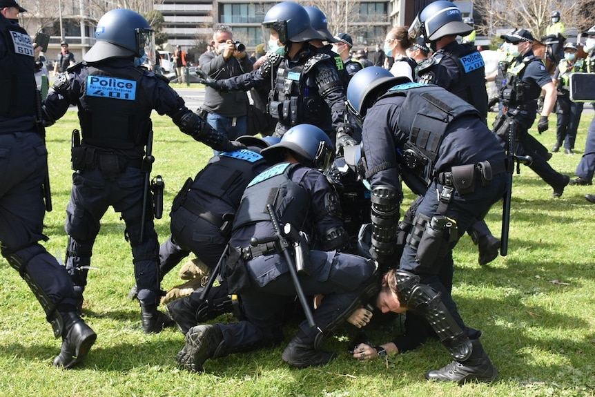 Police officers in helmets and padded jackets surround and kneel over a man as they arrest him at Albert Park Lake.