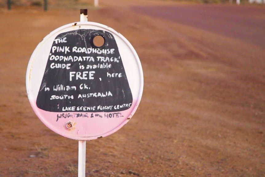 A small pink, black and white sign stands by the road describing the Oodnadatta Tracks guides available for free/