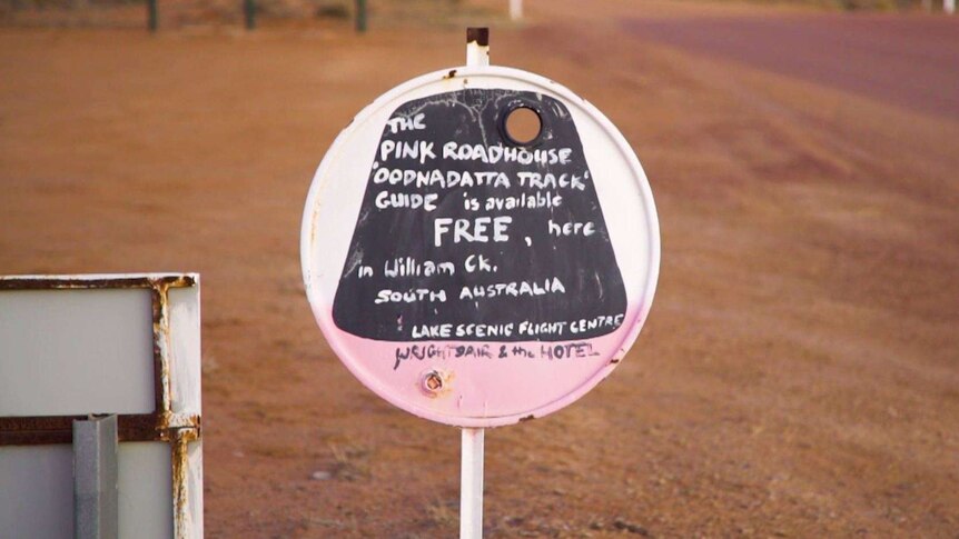 A small pink, black and white sign stands by the road describing the Oodnadatta Tracks guides available for free/