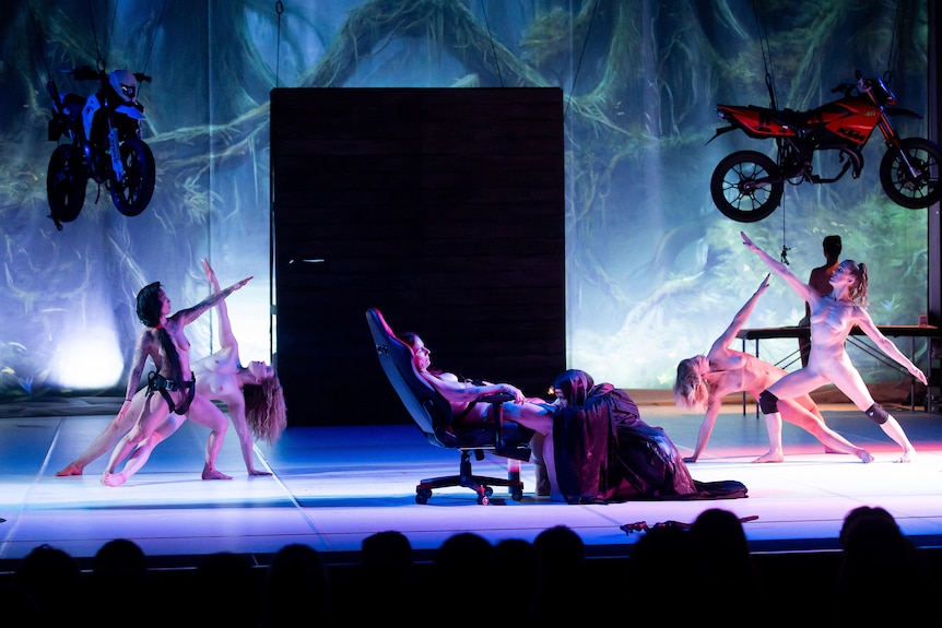 A group of six naked woman perform on stage, with motorbikes hanging above them.