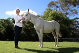 Sam Tassini (woman) smiles while patting a large white horse in a paddock.