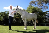 Sam Tassini (woman) smiles while patting a large white horse in a paddock.