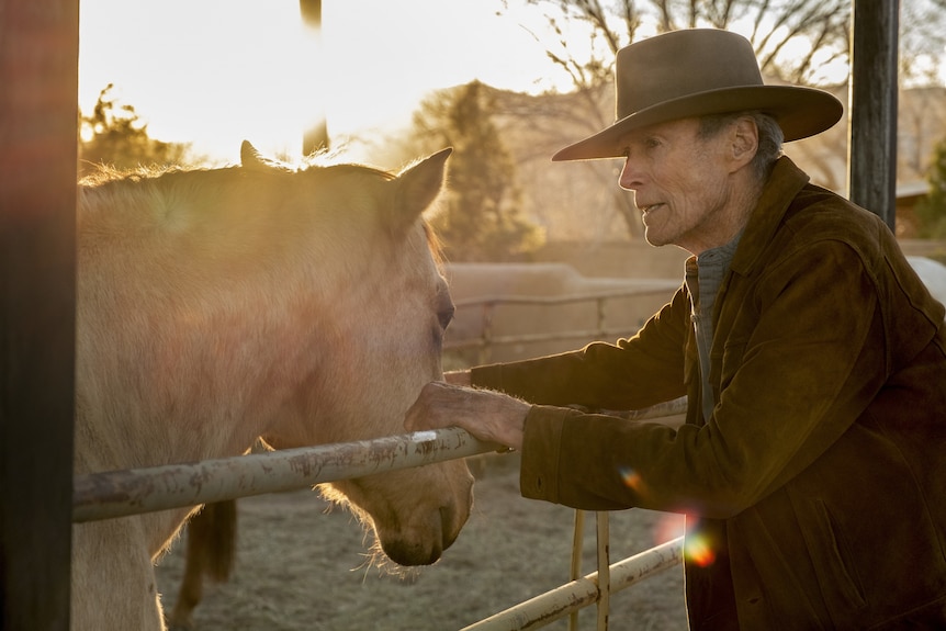 A contemplative 90-something man in a cowboy hat and jacket leans on a fence as he pats a horse