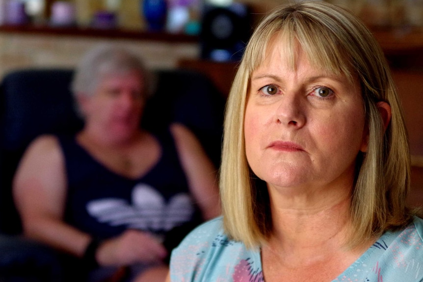 A woman looks at the camera with her husband in the background slumped in a chair.