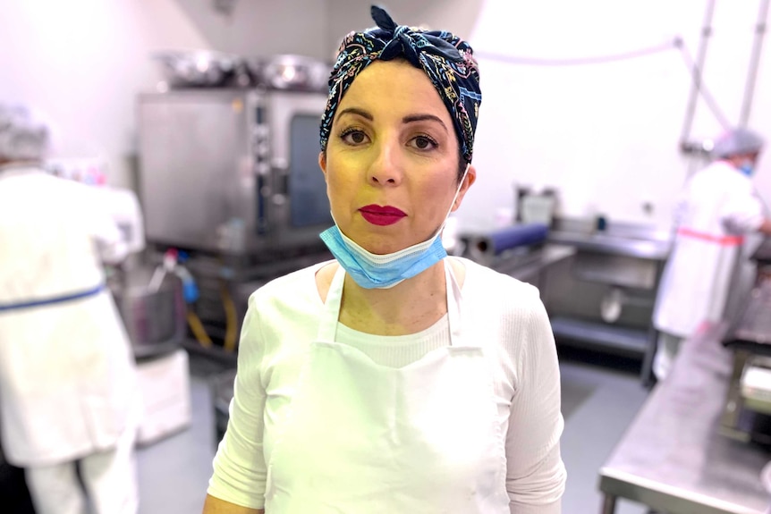A woman wearing a white apron and a black patterned headscarf, with a surgical mask pulled down.