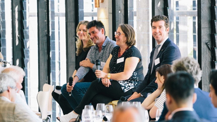 From left to right a female, male, female, male sit on stools before a crowd holding microphones presenting a panel talk on wine