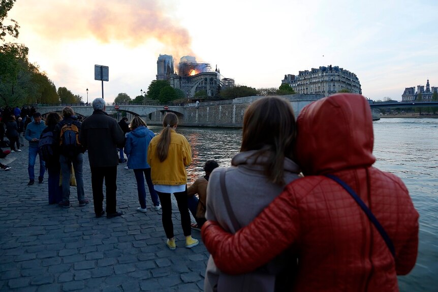 People gathered along a paved riverbank watch as flames and smoke rise from a burning catherdral in the distance