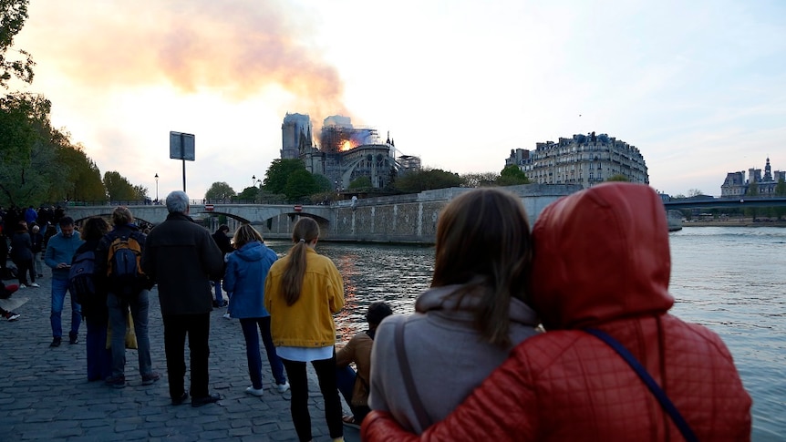 People gathered along a paved riverbank watch as flames and smoke rise from a burning cathedral in the distance