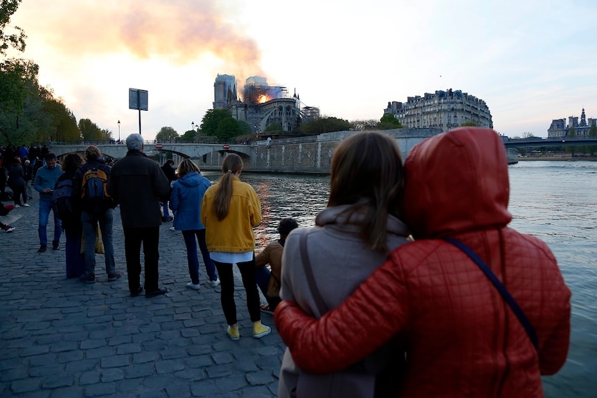 People gathered along a paved riverbank watch as flames and smoke rise from a burning cathedral in the distance