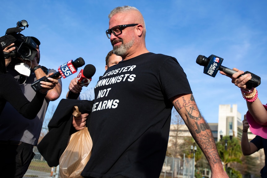 A man with tattoos wearing a black shirt and thick glasses and holding a yellow bag walking surrounded by news microphones