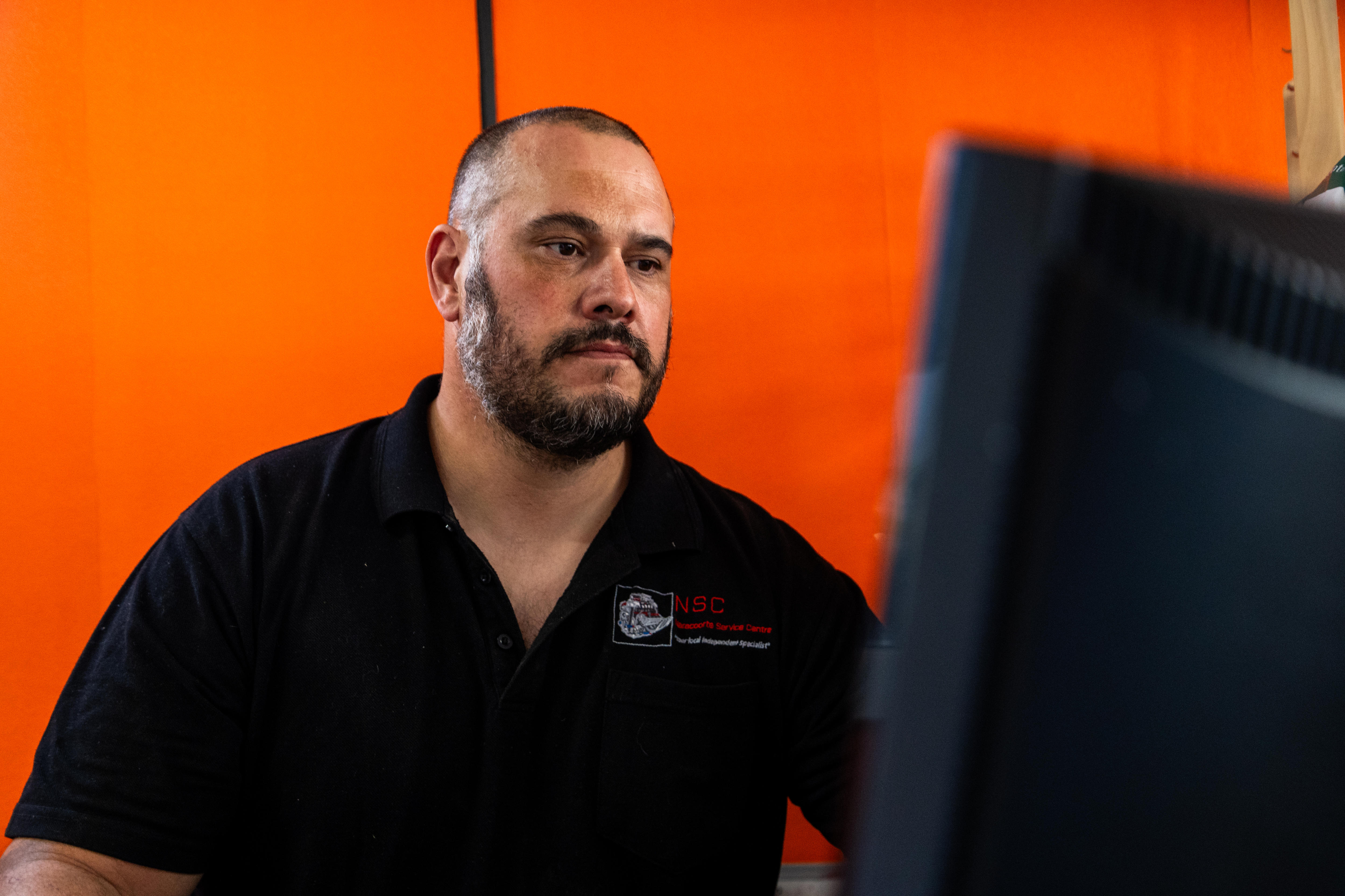 Um homem sentado em uma mesa em frente a uma parede laranja, com uma tela de computador à sua frente