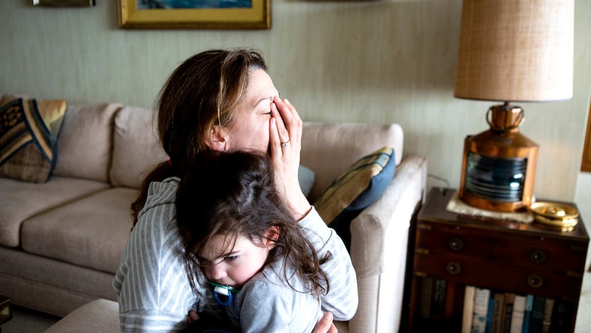 Tired mother and toddler hugging in living room. Mother has her hand over face.