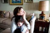 Tired mother and toddler hugging in living room. Mother has her hand over face.