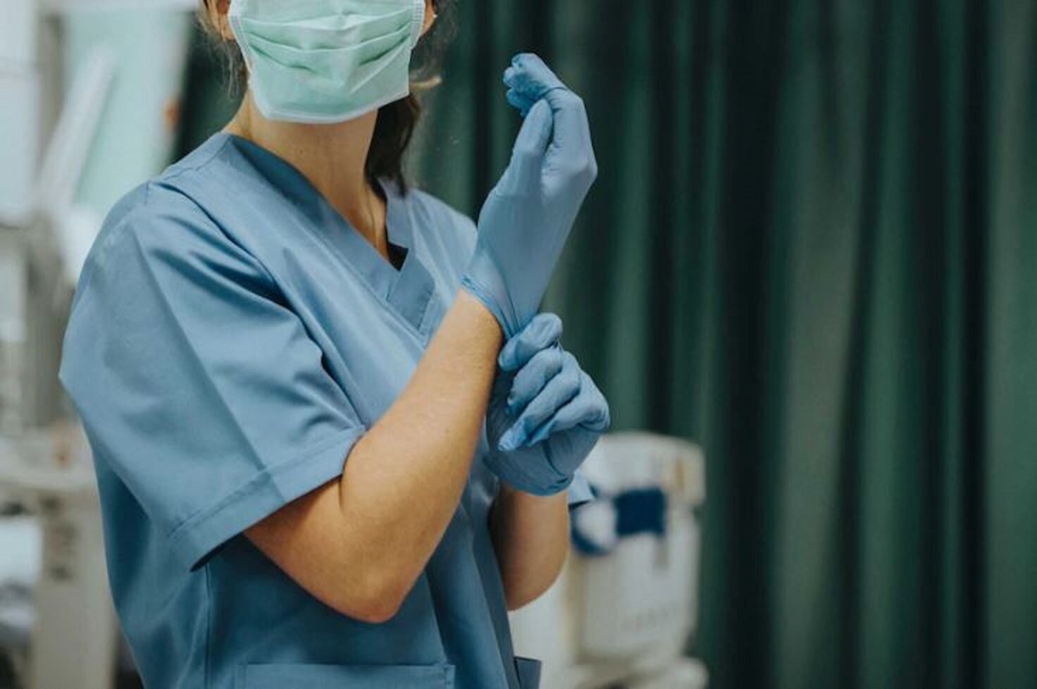 A nurse prepares for surgery wearing blue scrubs and a surgical mask and pulling on a pair of gloves in an operating theatre.