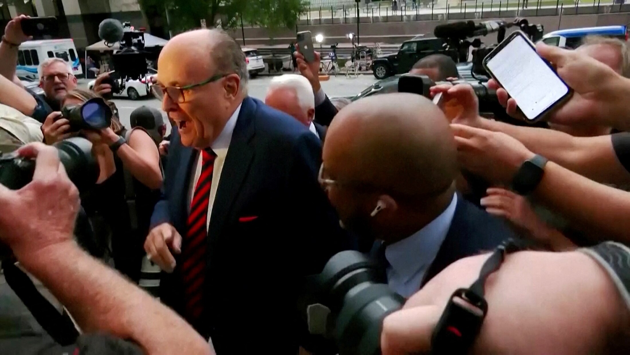 a bald man in glasses and a red tie pushes through a crowd of reporters