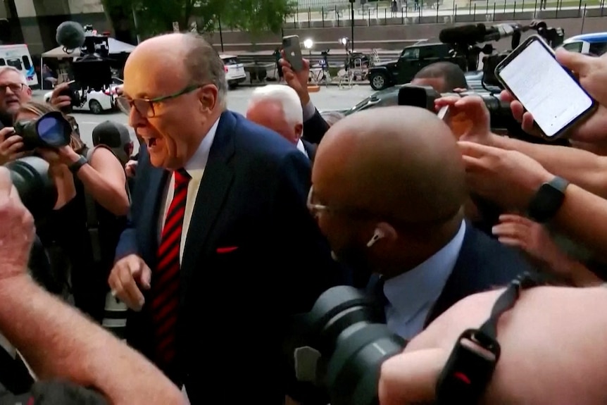 a bald man in glasses and a red tie pushes through a crowd of reporters