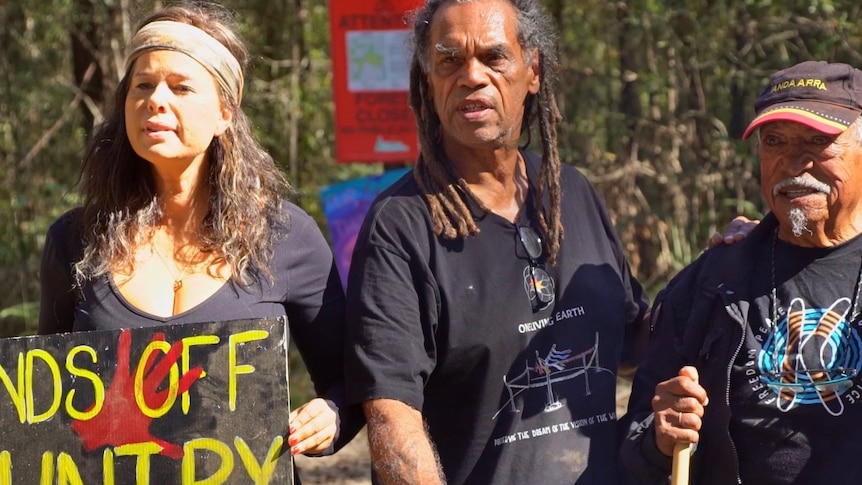 A woman and two men stand in a forest, holding anti-forestry signs.