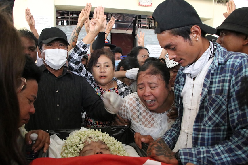 People crowd over coffin with dead body, woman cries as others raise three fingered salute.