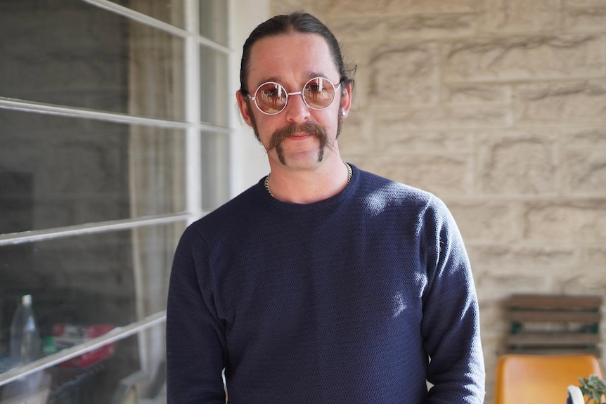 a man with glasses and handlebar moustache and dark top outside a house