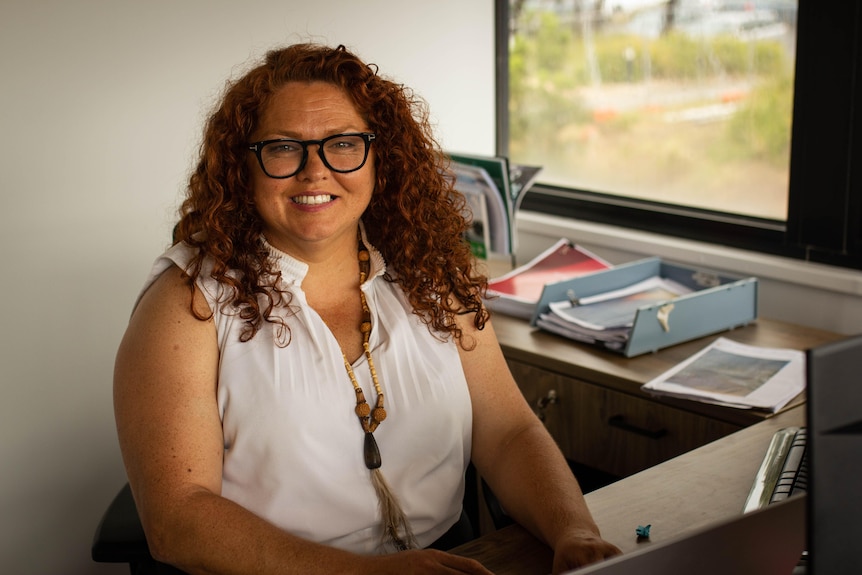 Portrait of a curly-haired woman with glasses sitting at a desk looking into the camera.