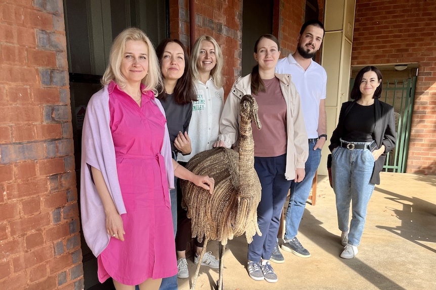 Six woman and a man stand outside a red brick building with an emu statue  