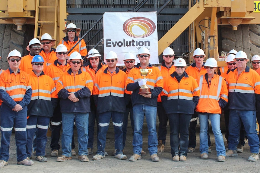 Mine workers in high visibility gear one in front of a big mining truck, one of them holding the Melbourne Cup