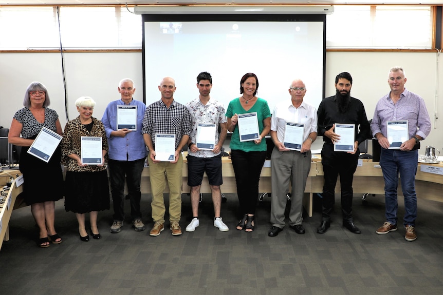 Nine people stand facing the camera holding up certificates showing their oaths and affirmations of office in local council