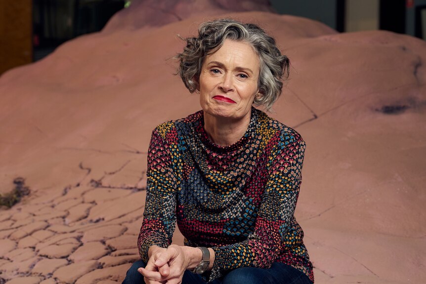 Judith Lucy, a middle-aged woman with curly, grey-streaked dark hair, sits on the edge of a barren landscape set, hands clasped