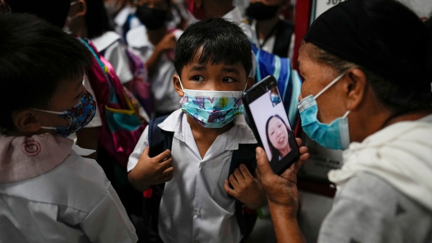 A boy wearing a mask on his first day of school talks to his mother on a smartphone video call.