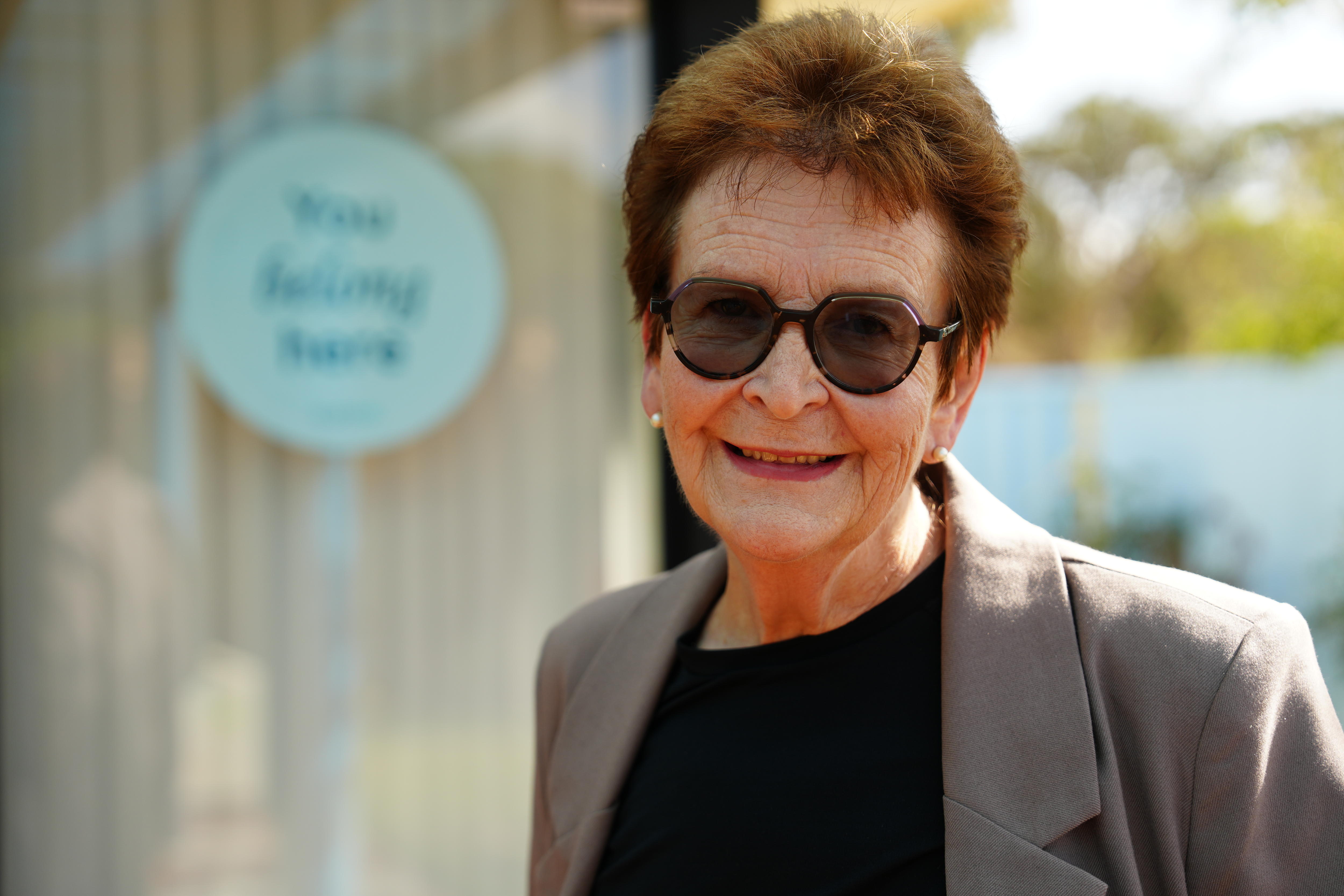 A head and shoulders shot of an older woman with brown hair, sunglasses, a grey jacket and black top, smiling outdoors.