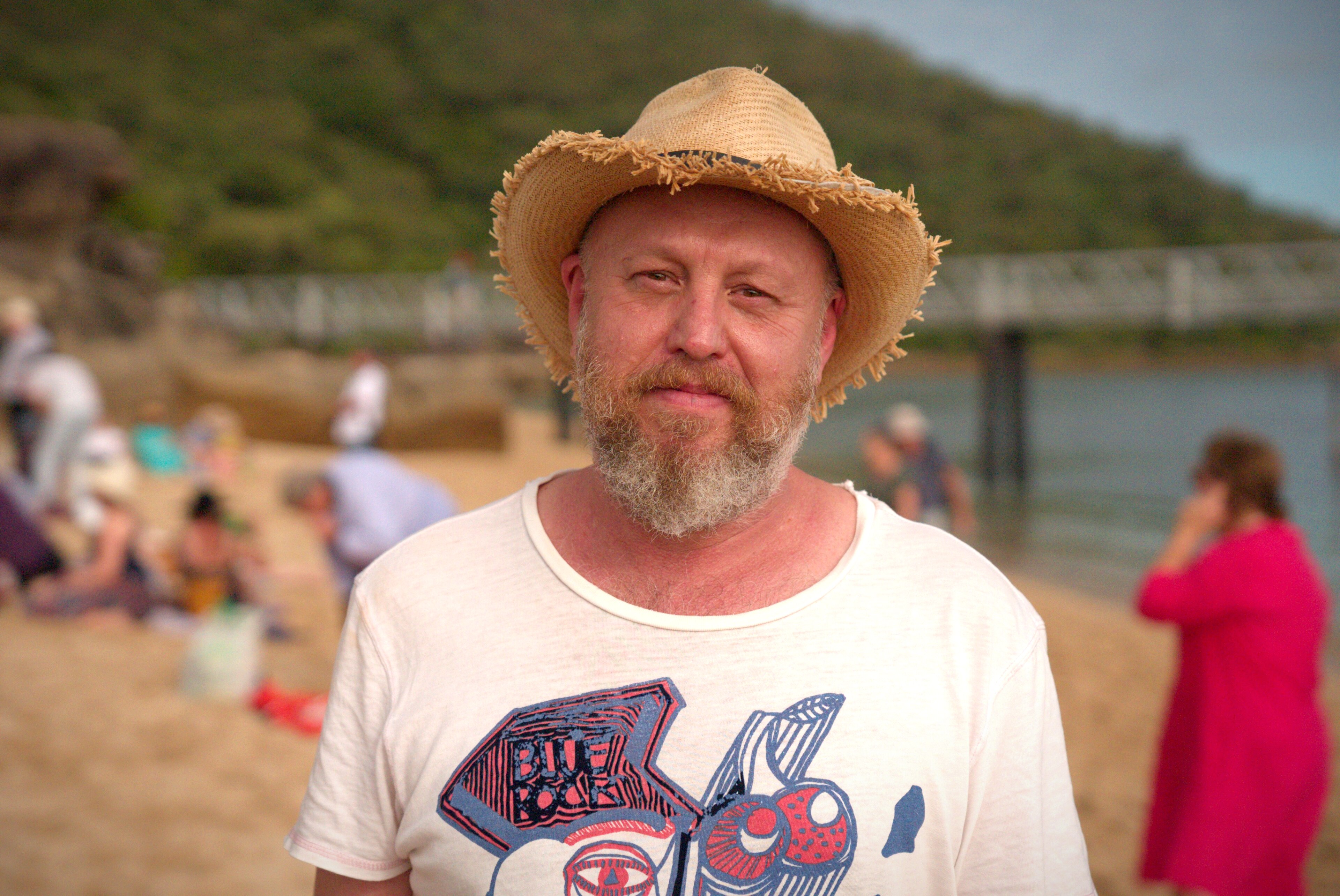 A man in a t-shirt and hat stands on a beach with crowds and a bridge in the background